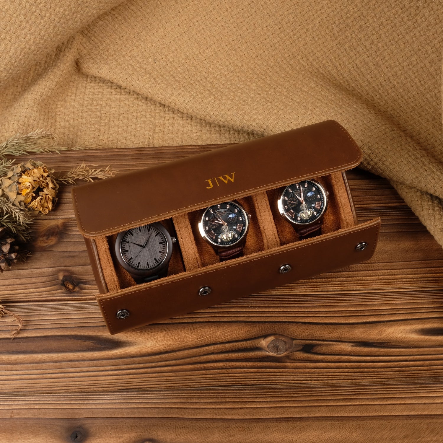 A brown leather watch case with multiple slots, each containing a watch, set against a wooden background with pine needles scattered around.