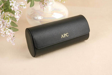 Black sunglasses case with 'APC' branding on a beige surface with floral elements.