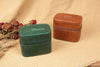 Two jewelry boxes, one green and one brown, on a textured surface with dried flowers.